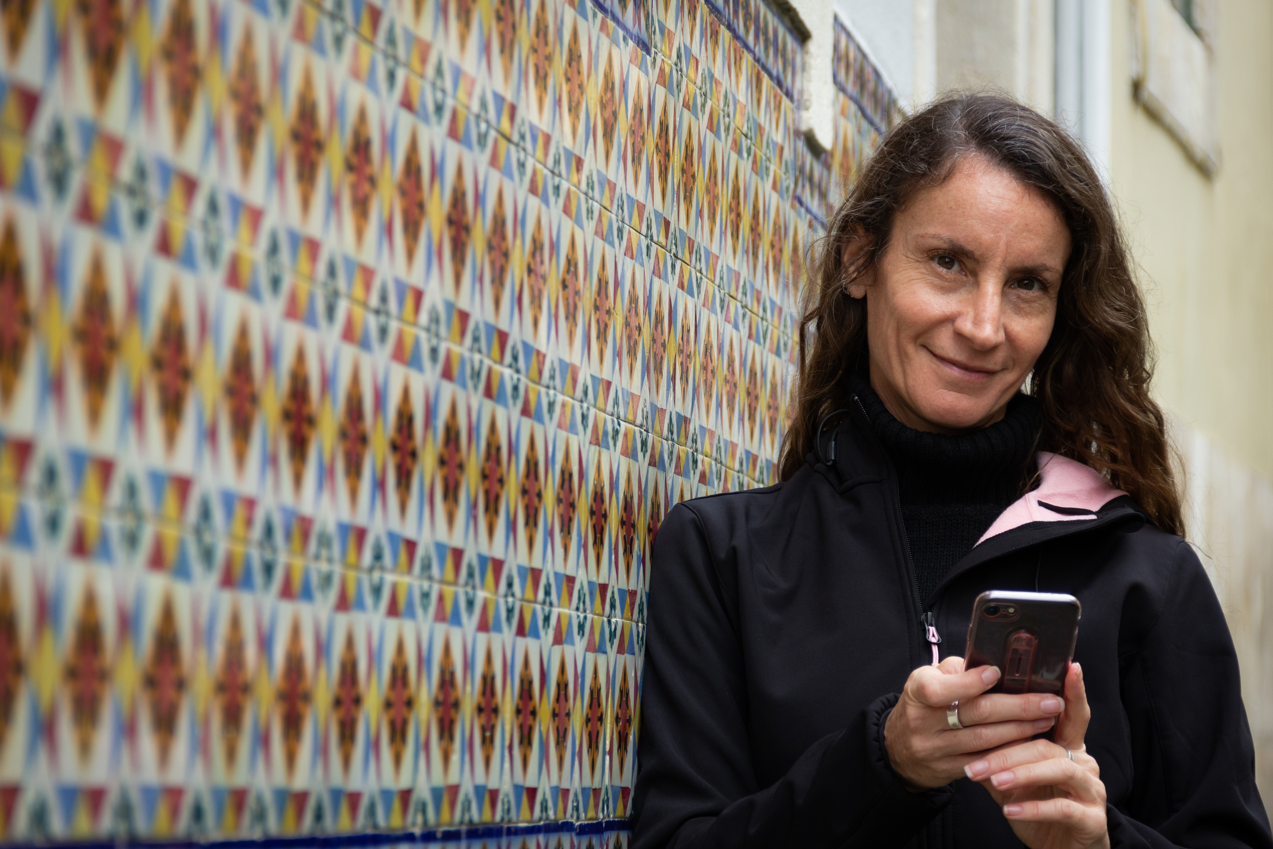 Smiling woman looking at camera holding cellphone and leaning on traditional tile wall building in Lisbon, Portugal. Connection, communication concepts Smiling woman looking at camera holding cellphone and leaning on traditional tile wall building in Lisbon, Portugal. Connection, communication concepts