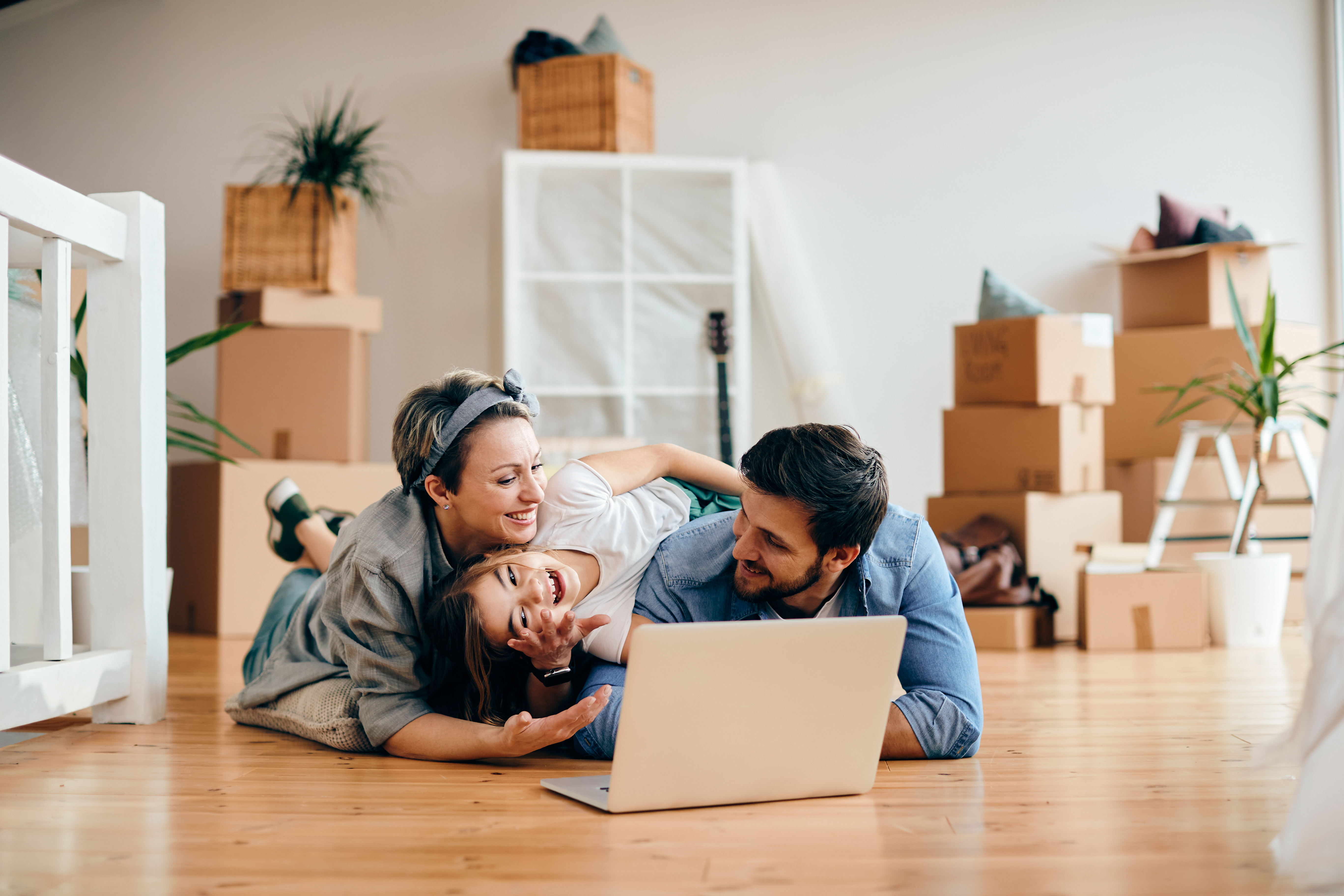 Cheerful family using laptop while relaxing on the floor at their new home. Happy parents and daughter having fun while using laptop and lying down on the floor after moving into new house.
