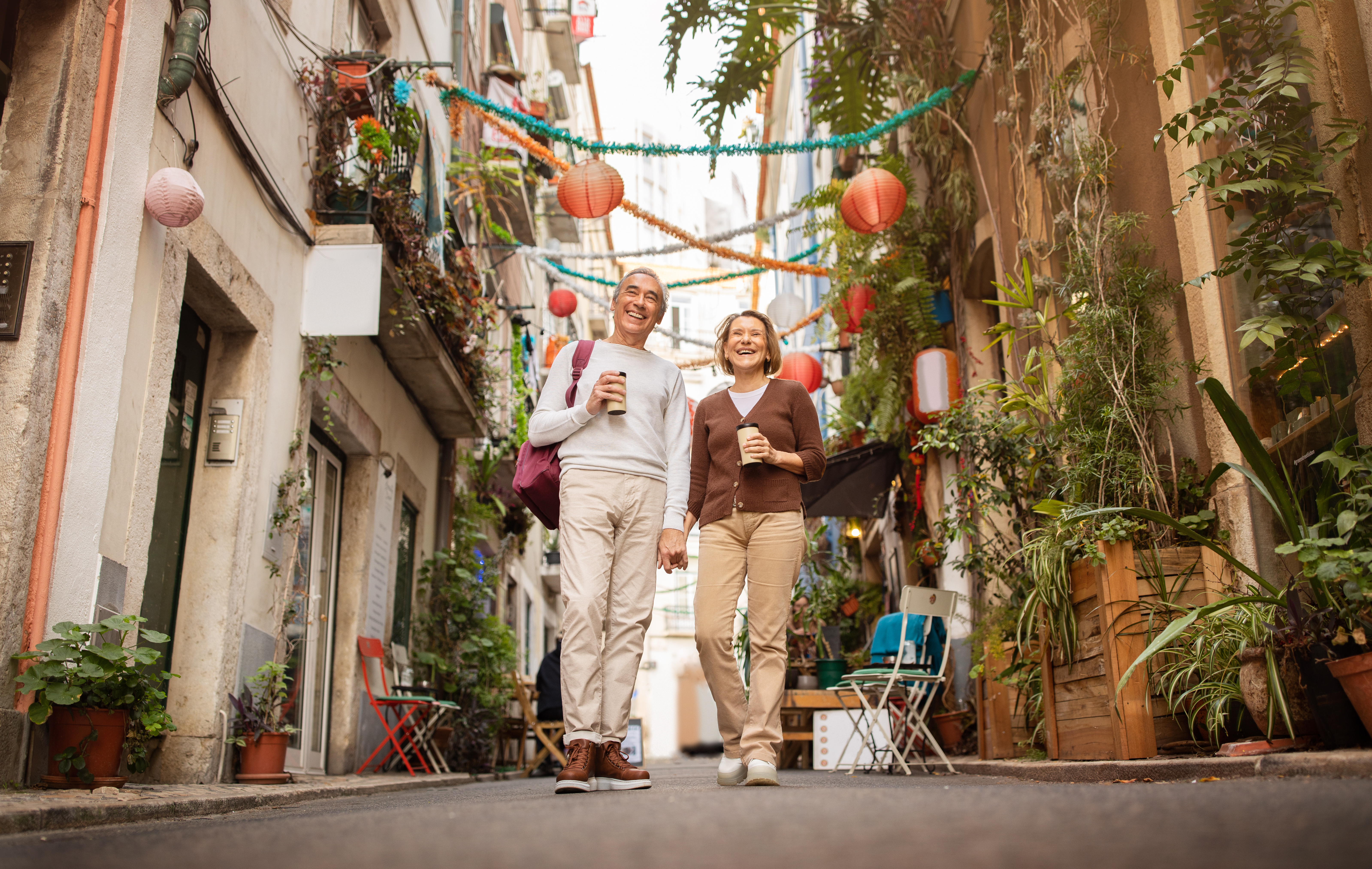 Cheerful Senior Travelers Couple Enjoying Walking In Lisbon Outside Cheerful Senior Travelers Couple Enjoying Walking Tour Holding Hands And Drinking Coffee On A Street In Lisbon Outside. Tourists Traveling In Europe After Retirement. Low Angle, Full Length Shot